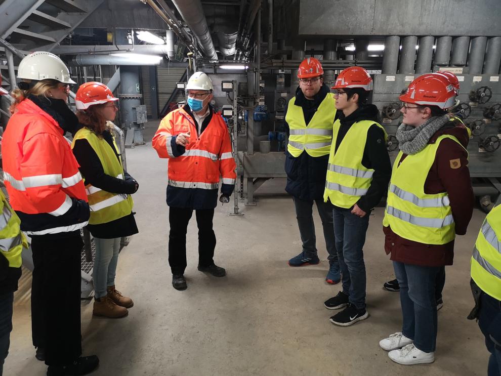 Eine Gruppe von Personen in Schutzausrüstung, einschließlich Schutzhelmen und Warnwesten, befindet sich im AEZ und erhält Erklärungen von Herrn Kellermann, der Betriebsleiter des AEZ.