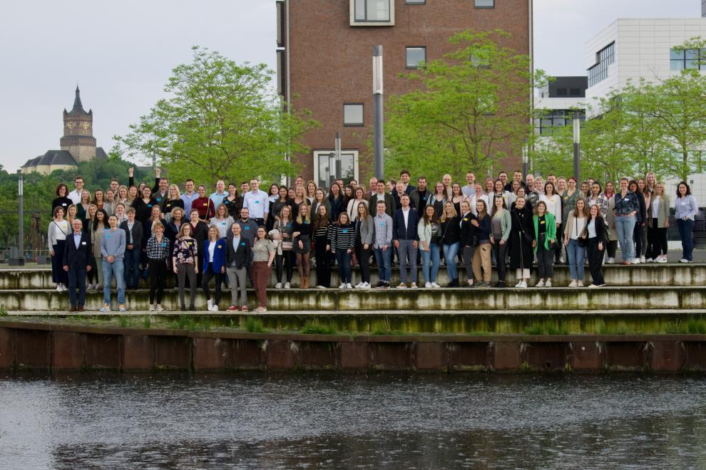 Eine große Gruppe von Alumni steht lächelnd vor einem Fluss, mit dem Campus im Hintergrund. Sie scheinen glücklich zu sein, sich wiederzusehen und genießen das Wiedersehen.