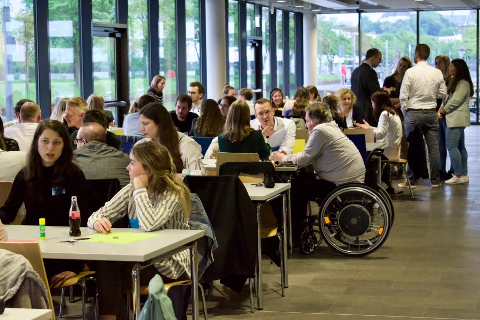 Alumni sitzen an Tischen in einem großen Raum mit Panoramafenstern und führen Gespräche. Einige Teilnehmer haben Getränke vor sich, und die Atmosphäre wirkt entspannt und freundlich.