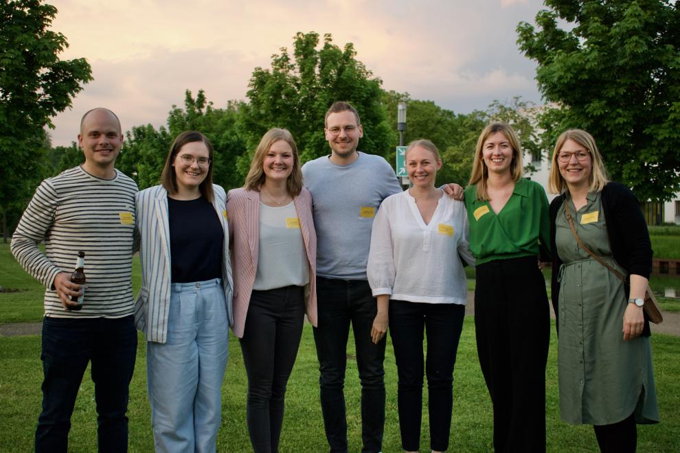 Eine kleine Gruppe von Alumni steht draußen im Grünen und lächelt in die Kamera. Sie halten Getränke in den Händen und genießen den geselligen Teil des Abends.