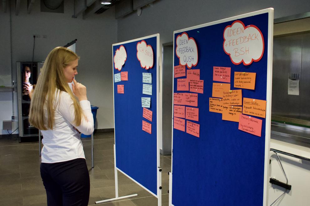 Eine junge Frau betrachtet nachdenklich eine Pinnwand mit vielen Notizen. Die Pinnwand ist in zwei Abschnitte unterteilt, "Ideen" und "Feedback", die jeweils für die Studiengänge QUSH und BSH stehen.