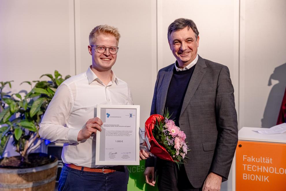 The image shows two men in an indoor setting. On the right stands Dr. Ralf Wimmer, recognizable by his smiling face and formal attire consisting of a gray suit and a dark turtleneck sweater. He is holding a bouquet of flowers in one hand and seems to be congratulating the man on the left. On the left stands Martin Willing, smiling broadly and holding a certificate symbolizing the award he has received. He is wearing a white shirt and has a more casual posture, with one hand holding the certificate. The certificate indicates an award of 1,000 euros and bears the logo of Rhine-Waal University of Applied Sciences and the Niederrhein Employers' Association.
