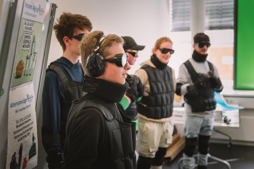 Fünf Schüler sind von der Seite zu sehen. Sie stehen in einer Reihe und blicken nach rechts in den Klassenraum. Alle tragen dunkle, seitlich geschlossene Brillen, Gehörschutz und gepolsterte Westen. Mit dieser Ausrüstung können die Schüler nachempfinden, wie es sich anfühlt, ein alter Mensch zu sein.