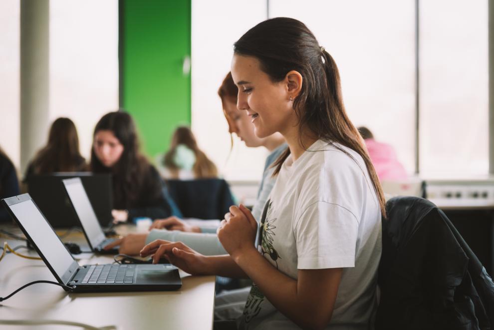 Im Vordergrund ist eine Schülerin in Seitenansicht zu sehen. Sie schaut glücklich auf den Bildschirm des Laptops, der vor ihr auf dem Tisch steht. Ihre Hand schwebt über der Tastatur. Ihr ist gerade etwas gelungen. Im Hintergrund arbeiten weitere Schülerinnen an ihren Laptops.