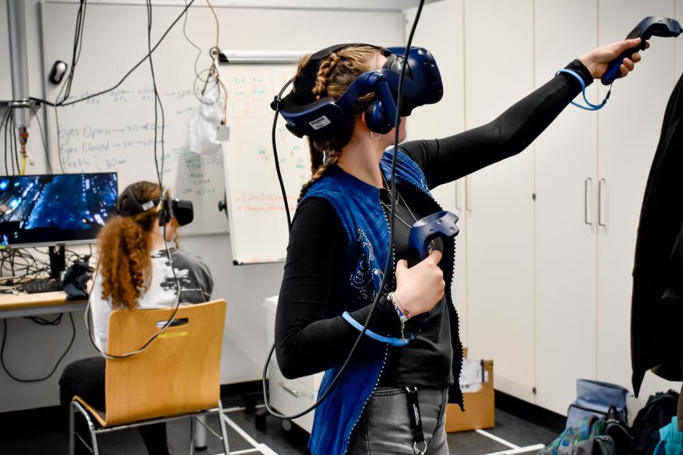 Im Vordergrund ist eine Schülerin zu sehen, die eine VR-Brille trägt und VR-Controller in den Händen hält Ihren rechten Arm hat sie vor ihrem Körper angewinkelt. Den linken Arm hat sie ausgestreckt und den Controller auf ein unsichtbares Ziel gerichtet.  Im Hintergrund sitzt eine weitere Schülerin mit VR-Brille auf einem Holzstuhl vor einem Monitor und schaut nach rechts ins Leere.