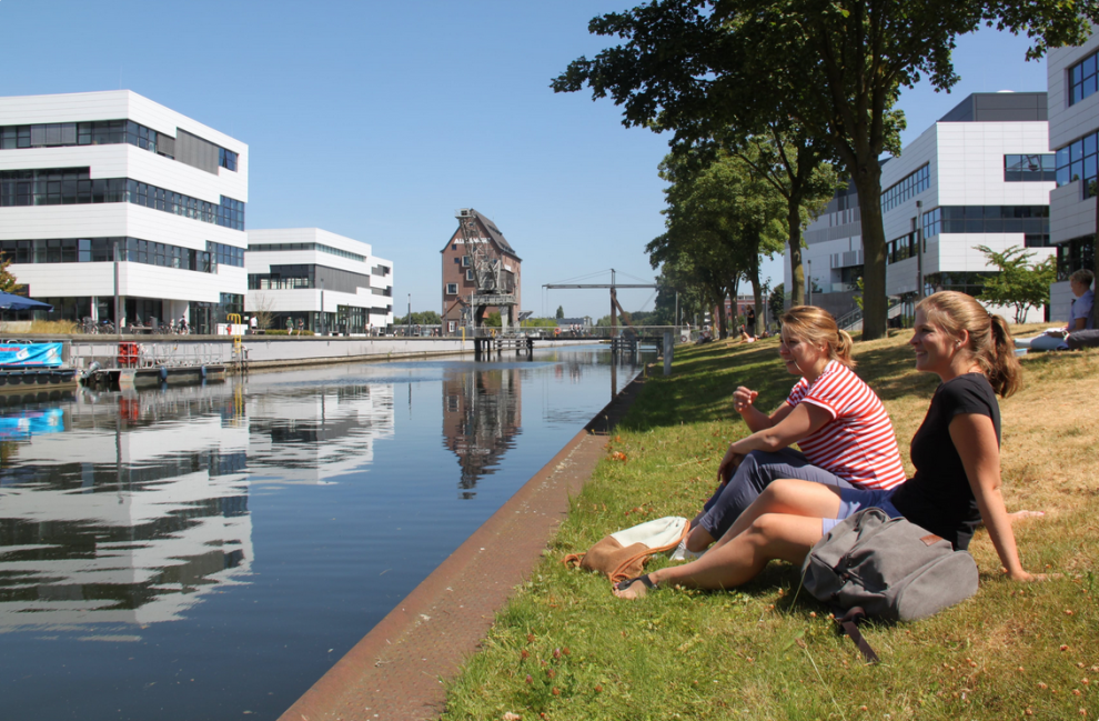 Zwei Frauen sitzen bei Sonnenschein auf einer Rasenfläche vor einem Fluss, im Hintergrund sind mehrere weiße Gebäude zu sehen.