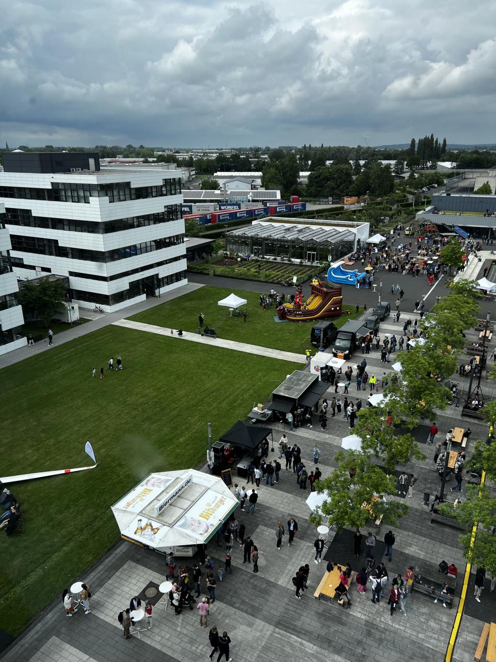 Eine Luftaufnahme des Campusfests, die das lebhafte Treiben auf dem Gelände zeigt. Viele Menschen sind auf dem Campus unterwegs, einige stehen an verschiedenen Ständen. Ein großes Gebäude und einige Gewächshäuser sind im Hintergrund zu sehen. Das Wetter ist leicht bewölkt.