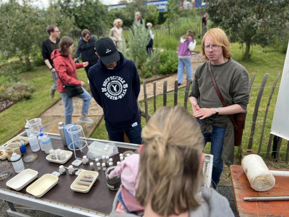 Ein Dozent erklärt den Besuchern verschiedene Bodenproben und Experimente. Im Hintergrund sieht man Besucher, die den Barfußpfad erkunden.