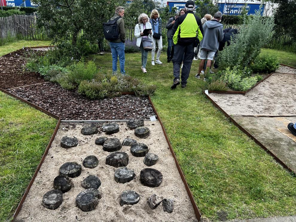 Besucher erkunden verschiedene Abschnitte des Lehrpfads, der unterschiedliche Bodenbeläge und Pflanzenarten zeigt. Ein Bereich des Pfads besteht aus Holzscheiben im Sand.