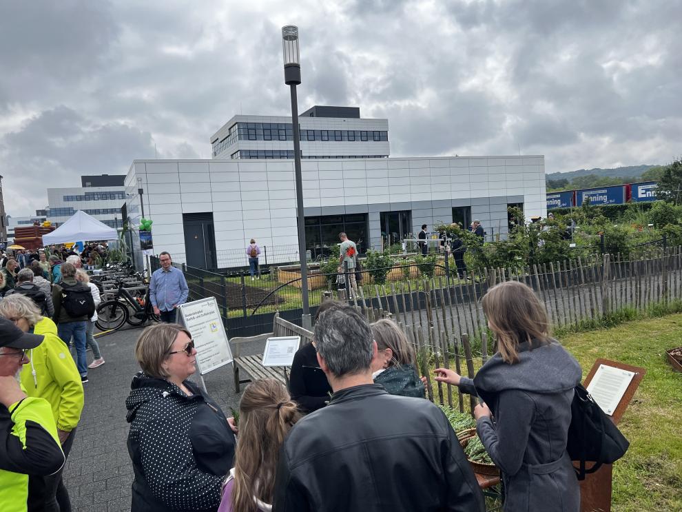 Eine Gruppe von Menschen steht um einen Informationsstand im Freien. Im Hintergrund sieht man den Barfuß- und Duftlehrpfad sowie die Gebäude der Hochschule.