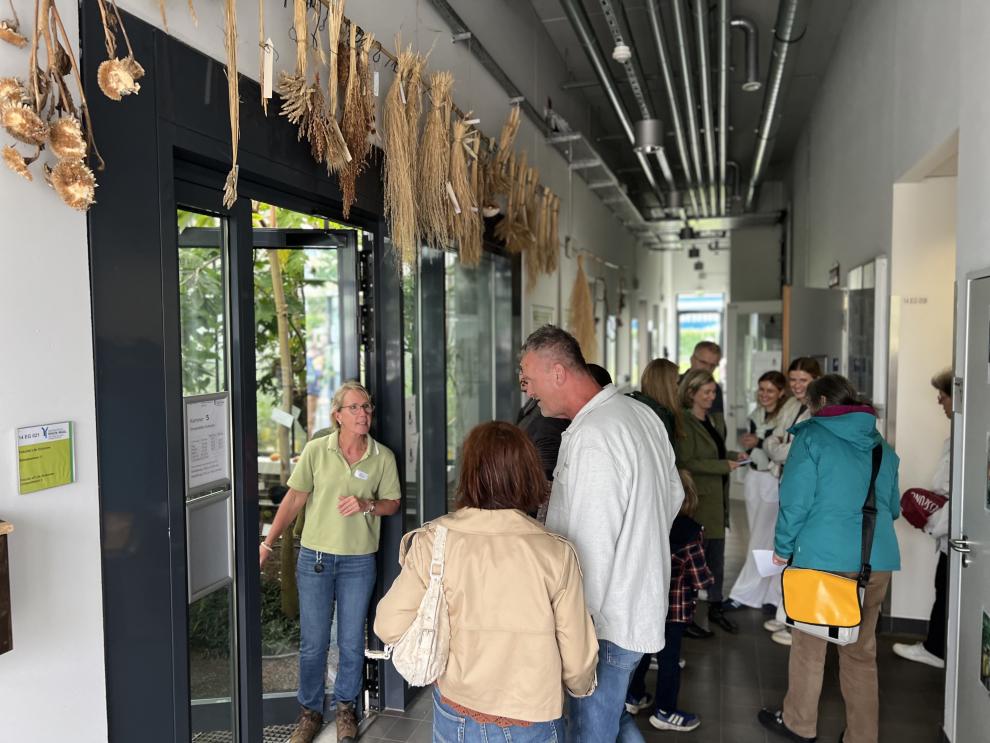 Eine Gruppe von Menschen steht im Flur des Tropenhaus, während eine Mitarbeiterin Informationen gibt. Der Eingang zum Tropenhaus ist im Hintergrund zu sehen.