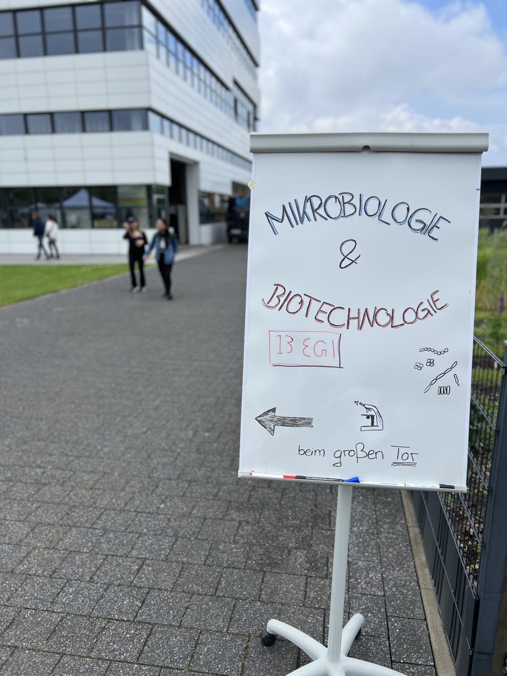 Ein Schild weist den Weg zur Ausstellung "Mikrobiologie & Biotechnologie" im Gebäude 13. Das Schild steht vor einem modernen Gebäude auf dem Campus.