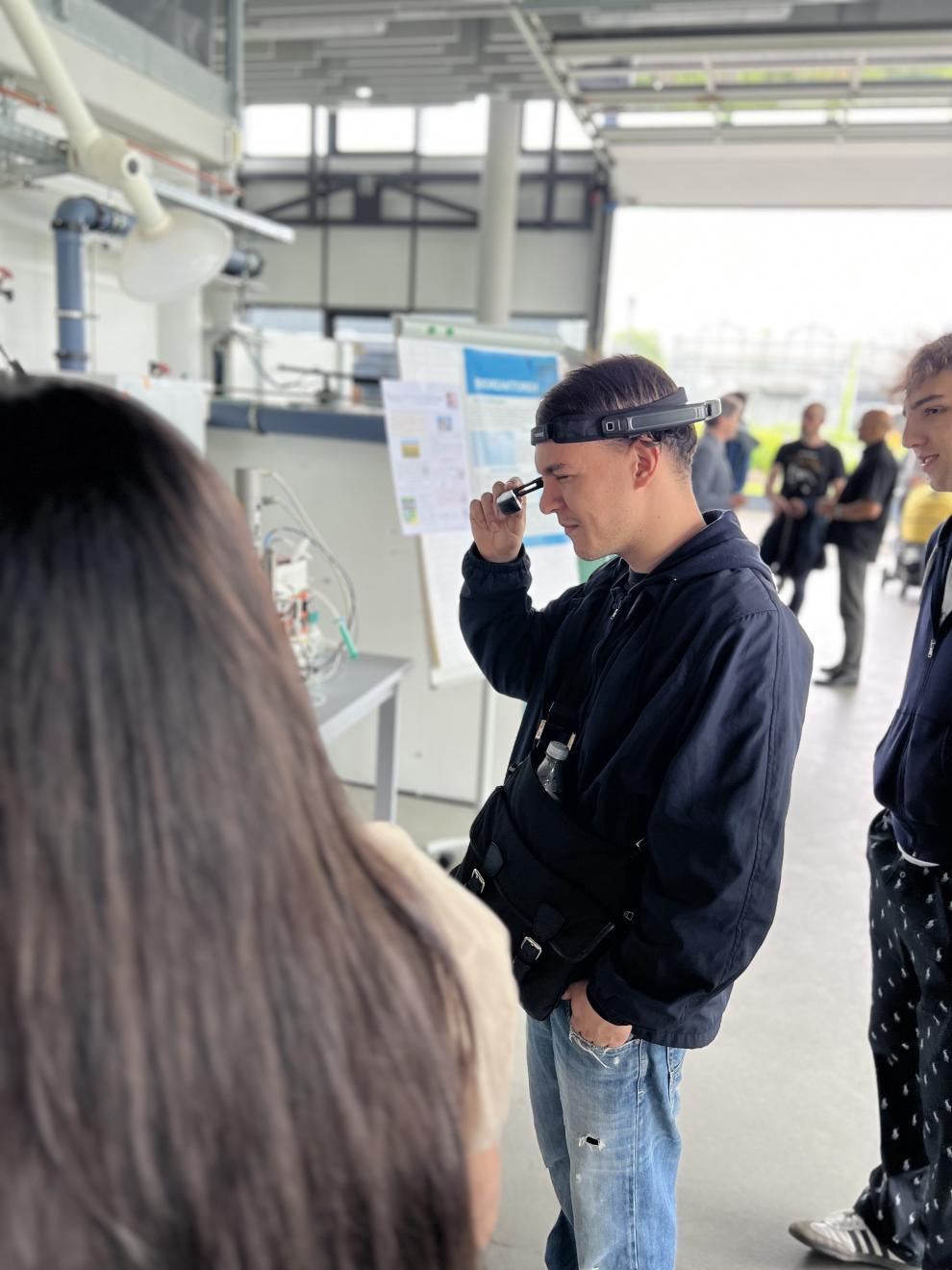 Ein junger Mann benutzt Smart Glasses während er sich eine Labor-Demonstration ansieht. Weitere Besucher stehen um ihn herum.