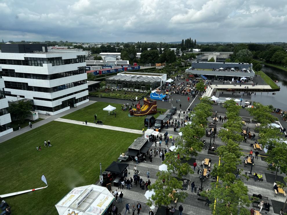 Eine weitere Luftaufnahme des Campusfests, die das Veranstaltungsgelände mit verschiedenen Ständen, einer Hüpfburg in Form eines Schiffs und vielen Besuchern zeigt. Im Hintergrund sind das Tropenhaus und Gebäude der Hochschule zu sehen.