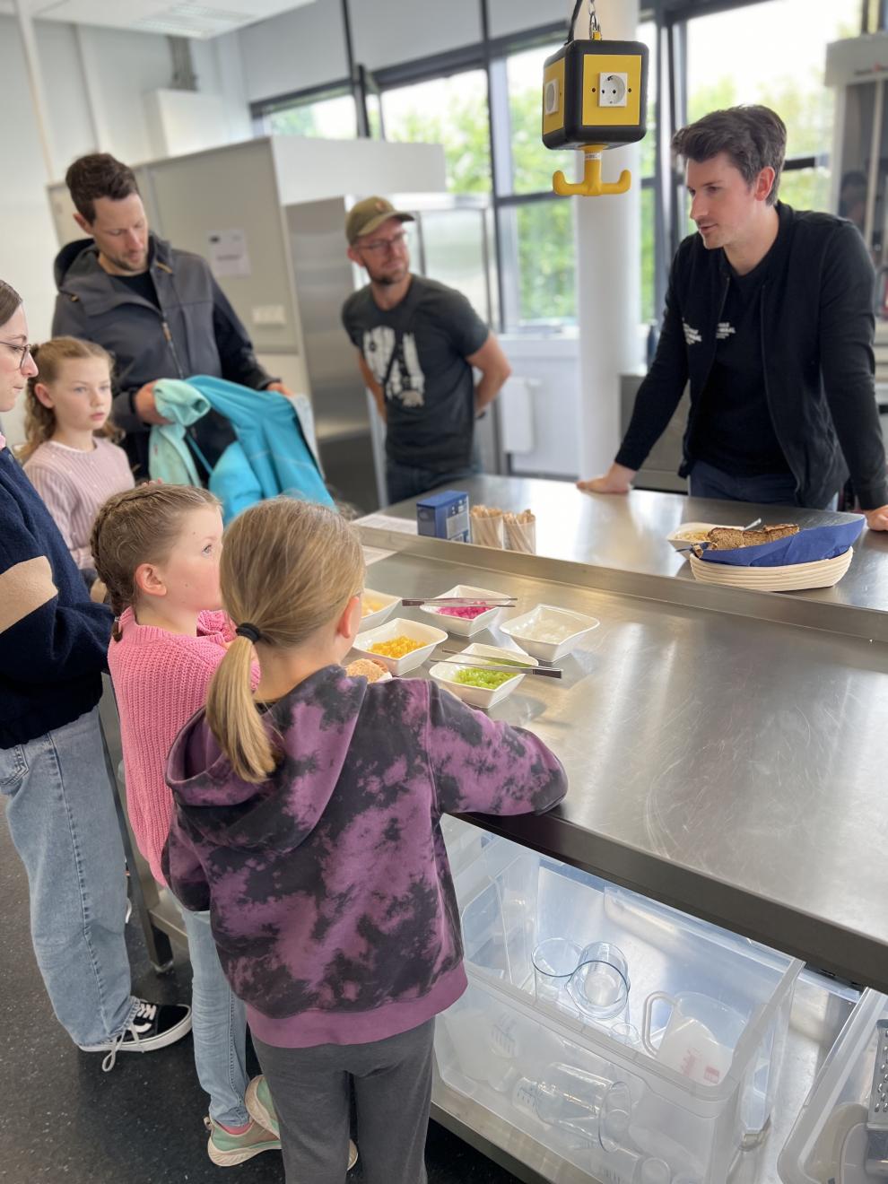 Kinder und Erwachsene stehen um eine Theke und betrachten die ausgestellten Lebensmittelproben. Ein wissenschaftlicher Mitarbeiter erklärt die verschiedenen Produkte und führt Experimente durch.