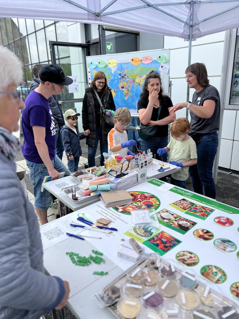 Zwei Mitarbeiter erklären Besuchern die gezeigten Materialien und Produkte an einem Informationsstand im Freien vor dem Tropenhaus. Verschiedene Poster und Proben sind auf dem Tisch ausgestellt.