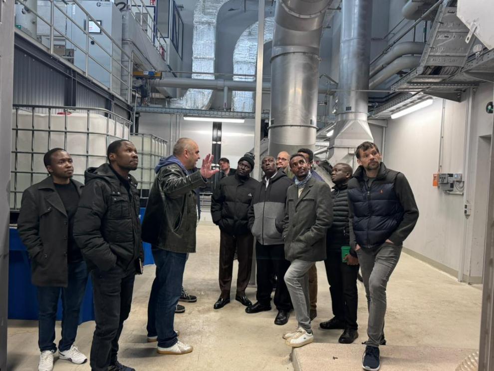 Mehrere Männer stehen draußen vor einem Gebäude, möglicherweise einem Bauernhof. Einer von ihnen, in einem lila T-Shirt, scheint etwas auf dem Boden zu erklären. Die Gruppe besteht aus Menschen verschiedener Hintergründe und sie stehen um ein Gerät herum.