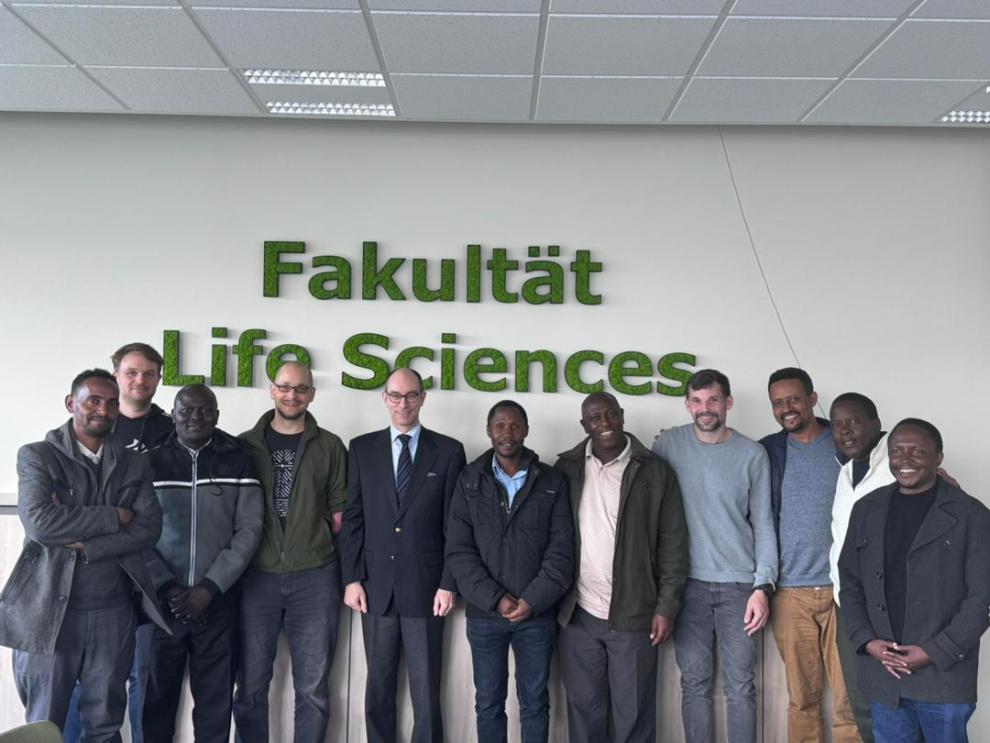 Eine Gruppe von Männern steht in einer industriellen Anlage. Ein Mann in einem schwarzen Lederjacke spricht und gestikuliert, während die anderen aufmerksam zuhören. Die Anlage scheint komplexe Rohrleitungen und Maschinen zu haben.