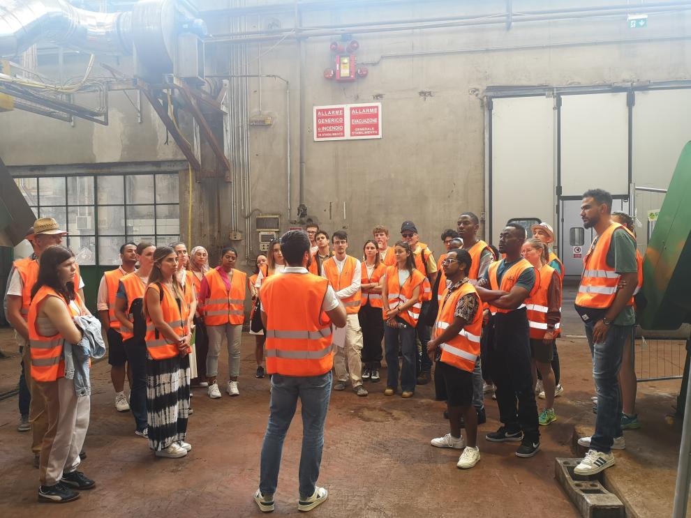 Studierende in orangefarbenen Sicherheitswesten hören einem Mitarbeiter in einer Produktionshalle zu. Dieses Bild ist während des Besuchs bei Caviro Company in Faenza entstanden sein, wo die Studierenden Einblicke in die Weinproduktion und Nachhaltigkeitsstrategien des Unternehmens erhielten.
