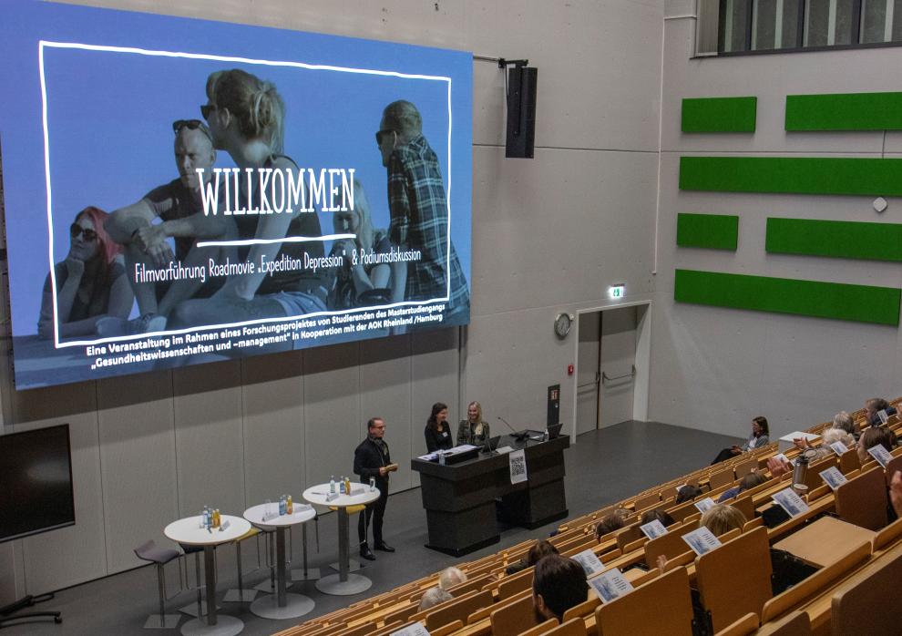 Filmvorführung des Roadmovies „Expedition Depression“. Im Vordergrund stehen drei Personen an einem Tisch mit Wasserflaschen, die sich auf die Veranstaltung vorbereiten oder moderieren. Der Raum ist mit Reihen von Stühlen ausgestattet, die teilweise besetzt sind, und es sind Informationsblätter auf den Sitzen ausgelegt. Die Wände sind hell und schlicht, mit einigen grünen Akzenten.