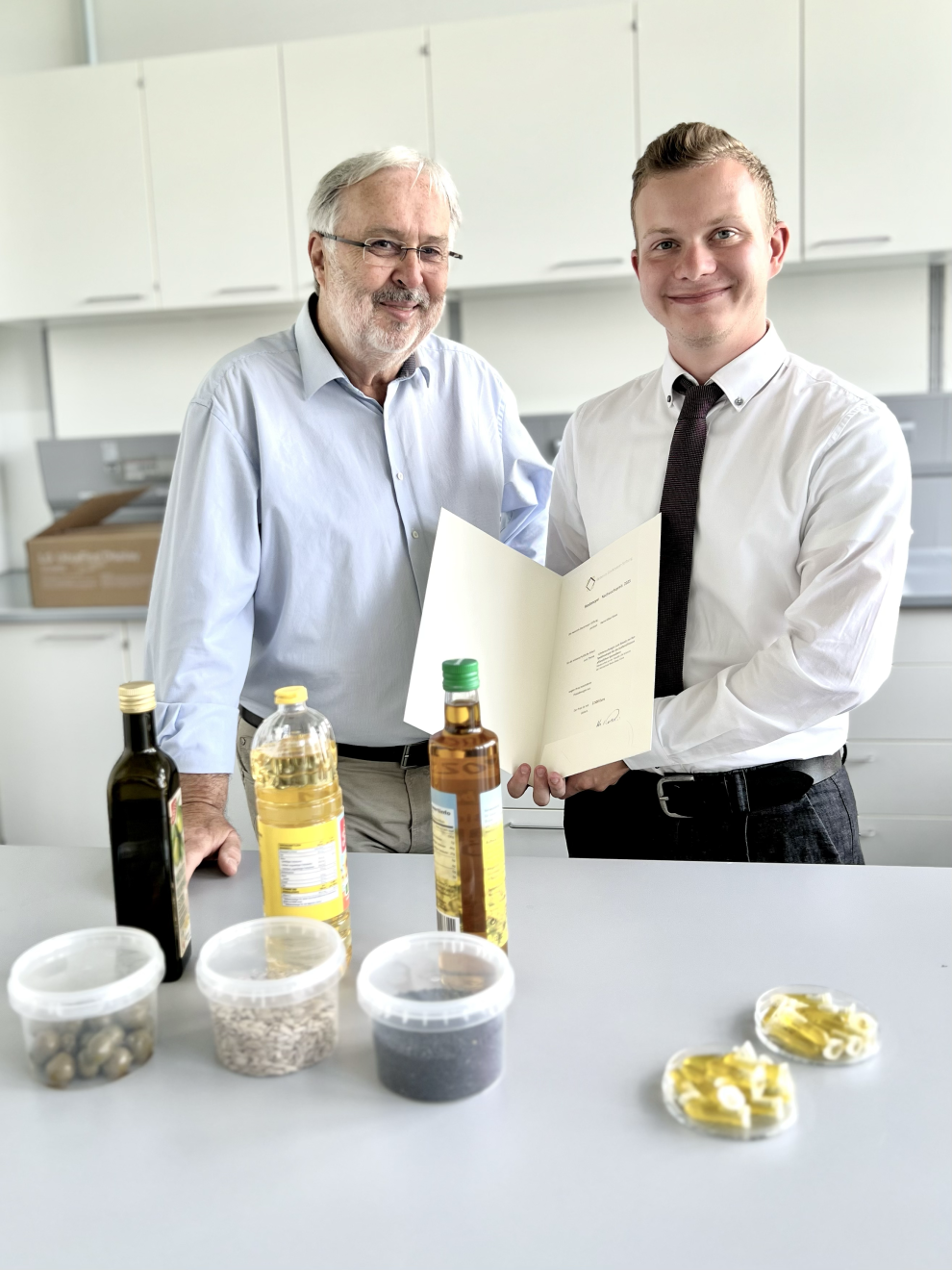 Ein jüngerer und ein älterer Mann präsentieren in einem Labor stolz eine Urkunde, während vor ihnen auf einem Tisch thematisch passende Produkte wie Speiseöle und Saaten ausgestellt sind.