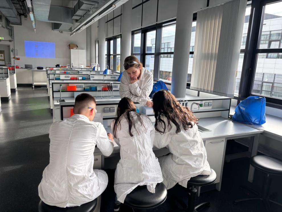 Eine Dozentin in Laborkittel und Schutzbrille beugt sich über einen Labortisch und betreut drei von hinten gezeigte Schülerinnen und Schüler. Die Gruppe sitzt auf Hockern und arbeitet konzentriert an einer praktischen Aufgabe.
