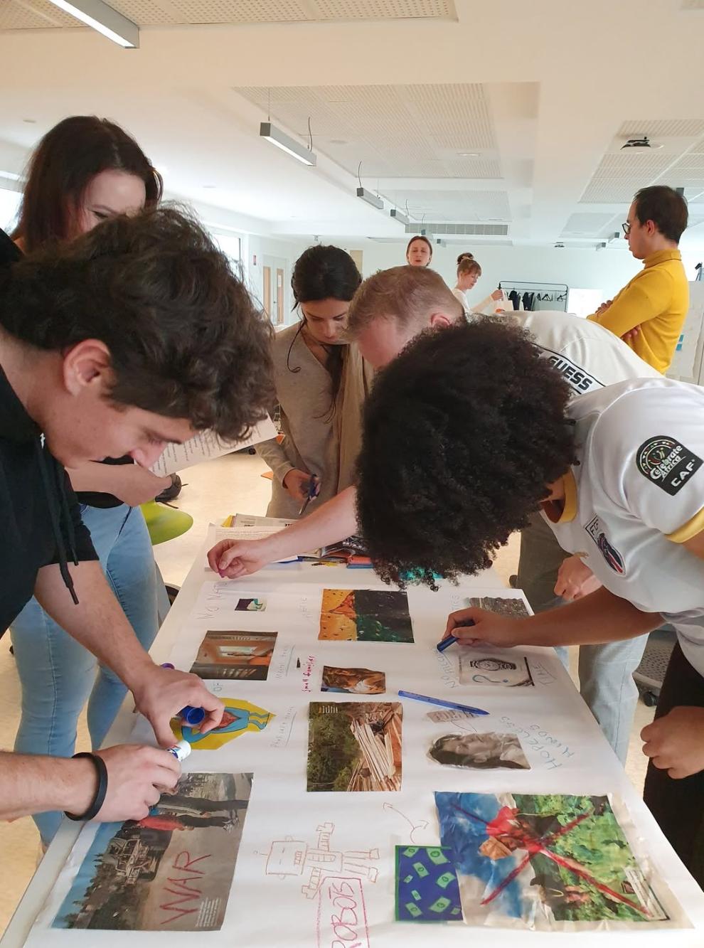 Mehrere Studierende beugen sich über einen großen weißen Tisch, auf dem Collagen aus Bildern und Texten liegen. Sie arbeiten konzentriert mit Stiften an ihren Projekten.