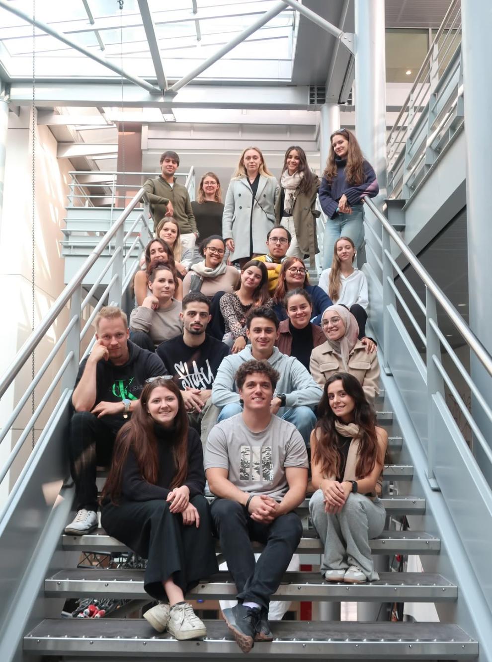 Eine große Gruppe junger, internationaler Studierender sitzt und steht lächelnd auf einer modernen Metalltreppe in einem Gebäude.