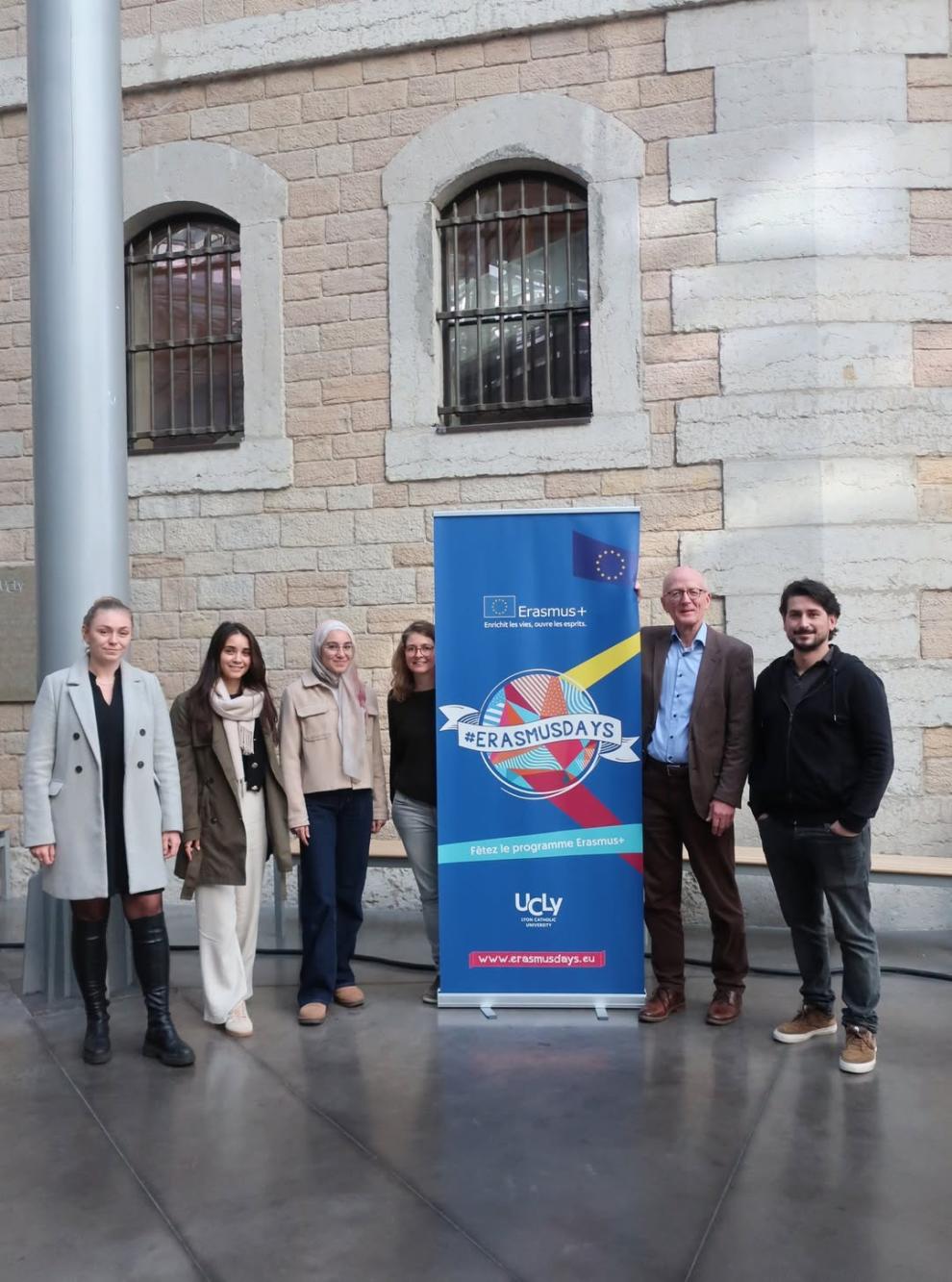 Eine Gruppe von sechs Personen, darunter Studierende und Lehrende, steht draußen vor einer alten Steinmauer neben einem blauen "Erasmus+ #ERASMUSDAYS" Banner.