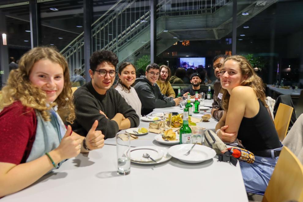 Eine Gruppe von Studierenden und Alumni sitzt lächelnd an einem Tisch mit Getränken und Snacks; eine Person zeigt einen Daumen nach oben in die Kamera.