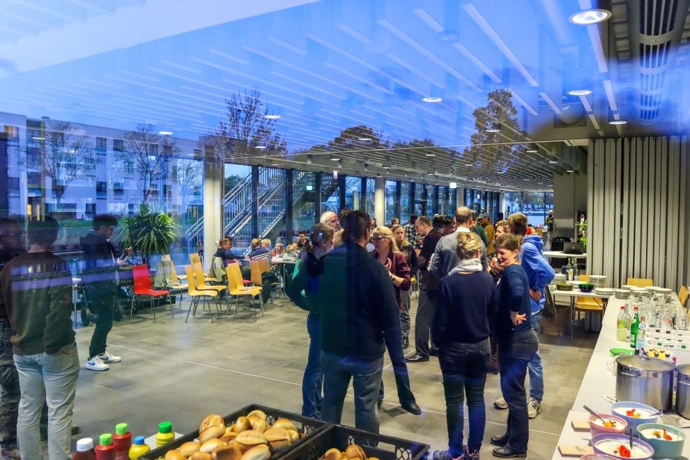 Ein gut gefüllter Veranstaltungsraum mit großen Fensterfronten, in dem Gäste stehen und sich unterhalten; im Vordergrund und rechts ist ein Buffet mit Speisen und Getränken aufgebaut.