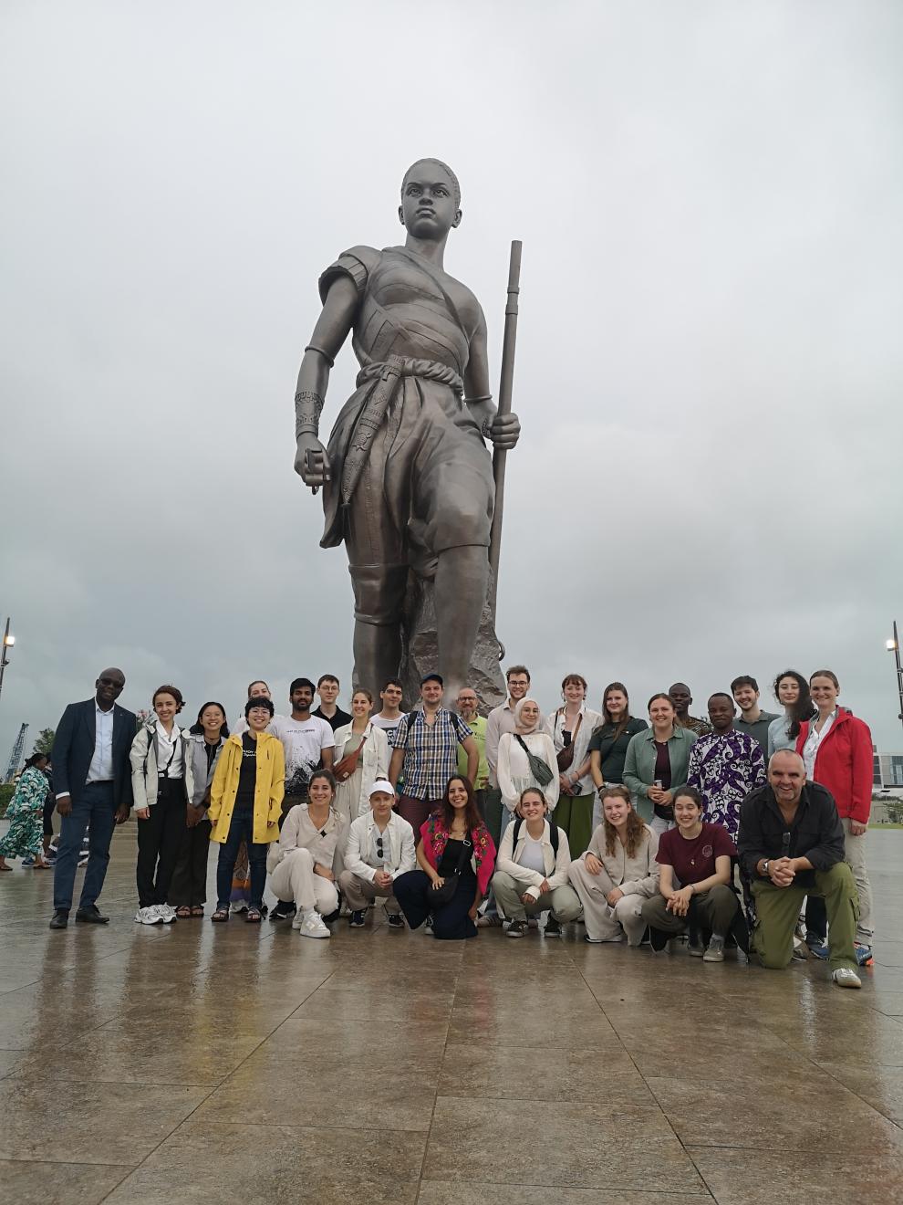Gruppenfoto der Studierenden und Professoren vor der monumentalen, bronzenen Amazonen-Statue in Cotonou an einem regnerischen Tag. Die riesige Figur einer Kriegerin ragt im Hintergrund in den Himmel.