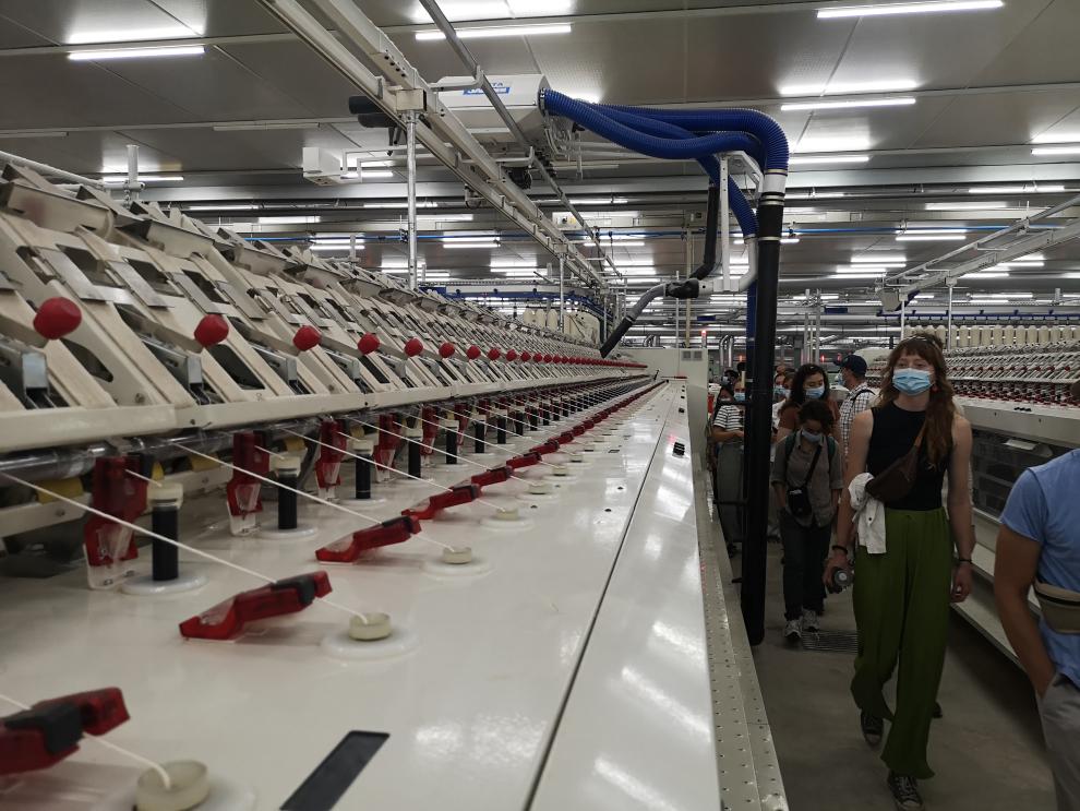 Blick in eine große, moderne Industriehalle der Textilproduktion. Man sieht lange Reihen von weißen Spinnereimaschinen, an denen die Besuchergruppe in einem Gang entlangläuft.