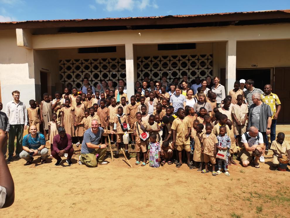 Ein fröhliches Gruppenbild vor einem Schulgebäude. Die Studierenden der Hochschule haben sich mit Dutzenden Schulkindern in beigen Uniformen gemischt; einige knien vorne, alle schauen in die Kamera.
