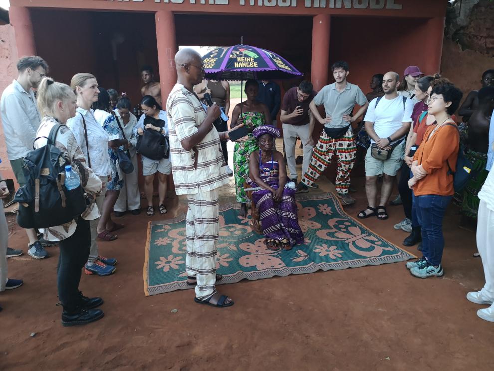 Eine traditionelle Zeremonie in einem Innenhof. Ein Mann in gemusterter Kleidung spricht in ein Mikrofon, gerichtet an eine Würdenträgerin, die unter einem bunten Sonnenschirm sitzt, während die Studierenden zuhören.