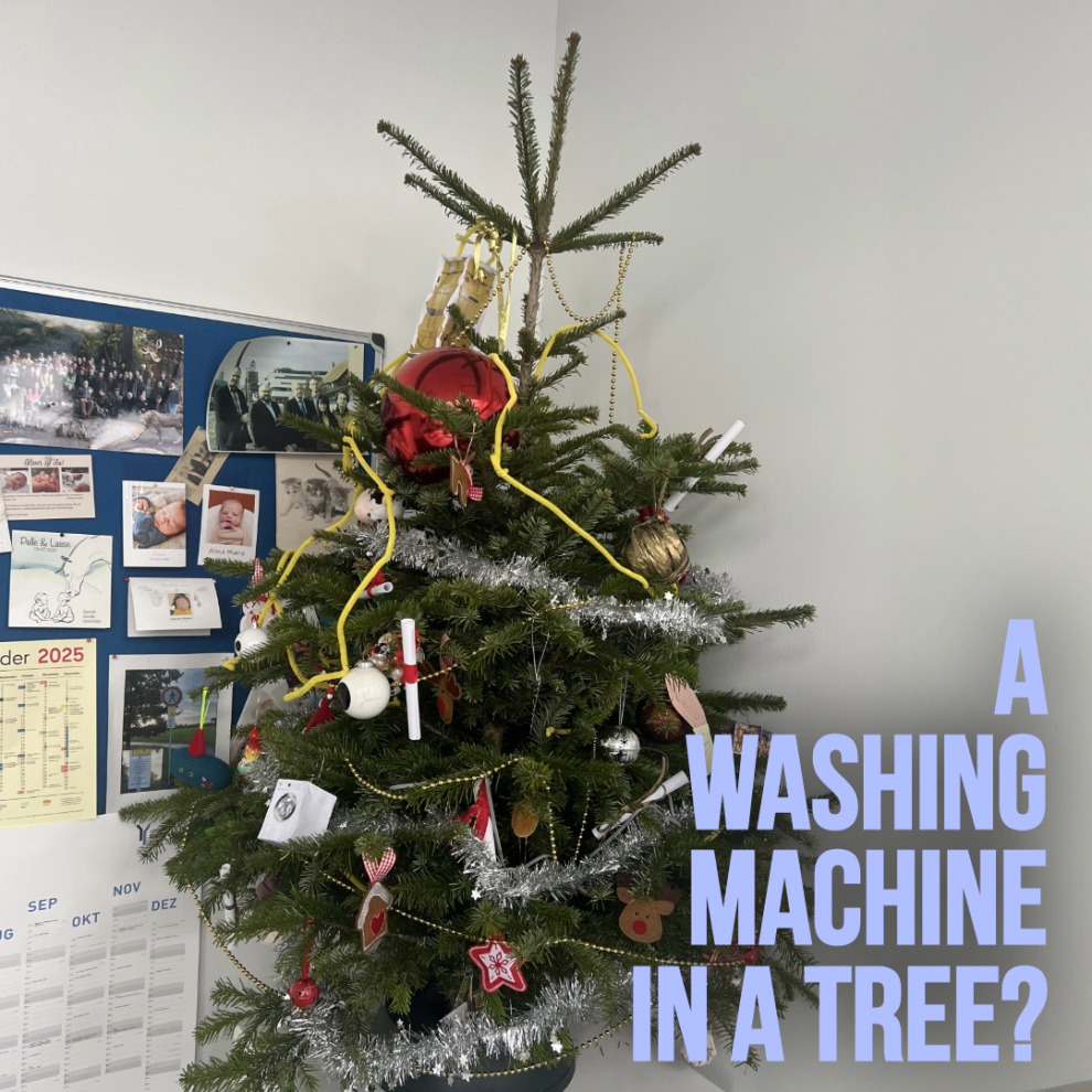 Weihnachtsbaum im Büroraum, geschmückt mit wissenschaftlichen Objekten wie Reagenzgläsern, gelben Laborschläuchen und Papieranhängern in Form von Waschmaschinen.