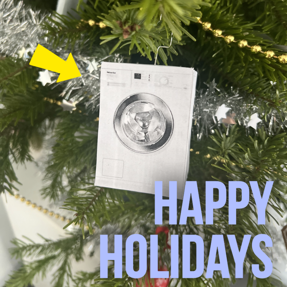 Nahaufnahme eines Weihnachtsbaumzweiges mit einem Papieranhänger, der eine moderne weiße Waschmaschine zeigt, umgeben von silbernem Lametta.