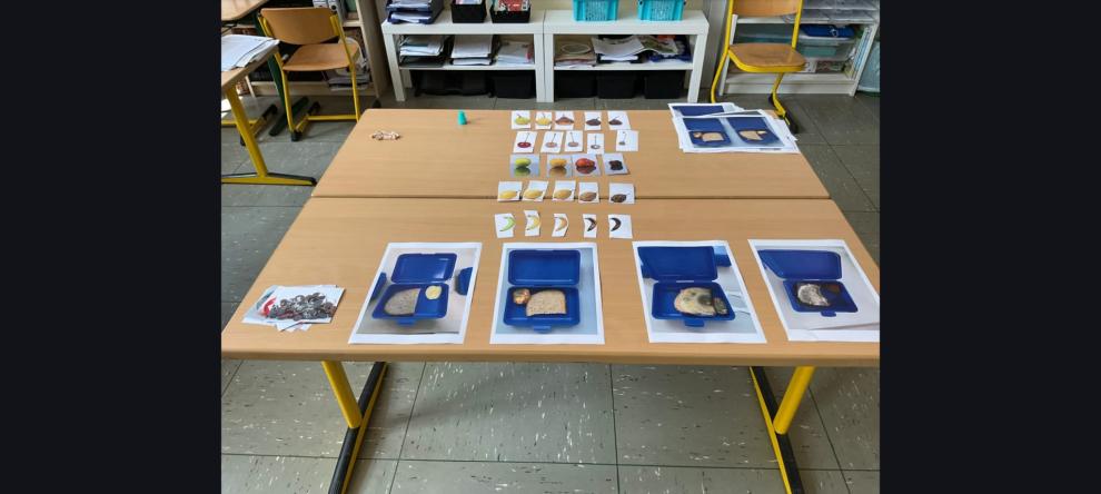 Zwei zusammengeschobene Tische in einem Klassenzimmer. Darauf sind anschauliche Bildkarten für ein Experiment ausgelegt, die in mehreren Stufen zeigen, wie Obst verdirbt und Pausenbrote in blauen Brotdosen verschimmeln.