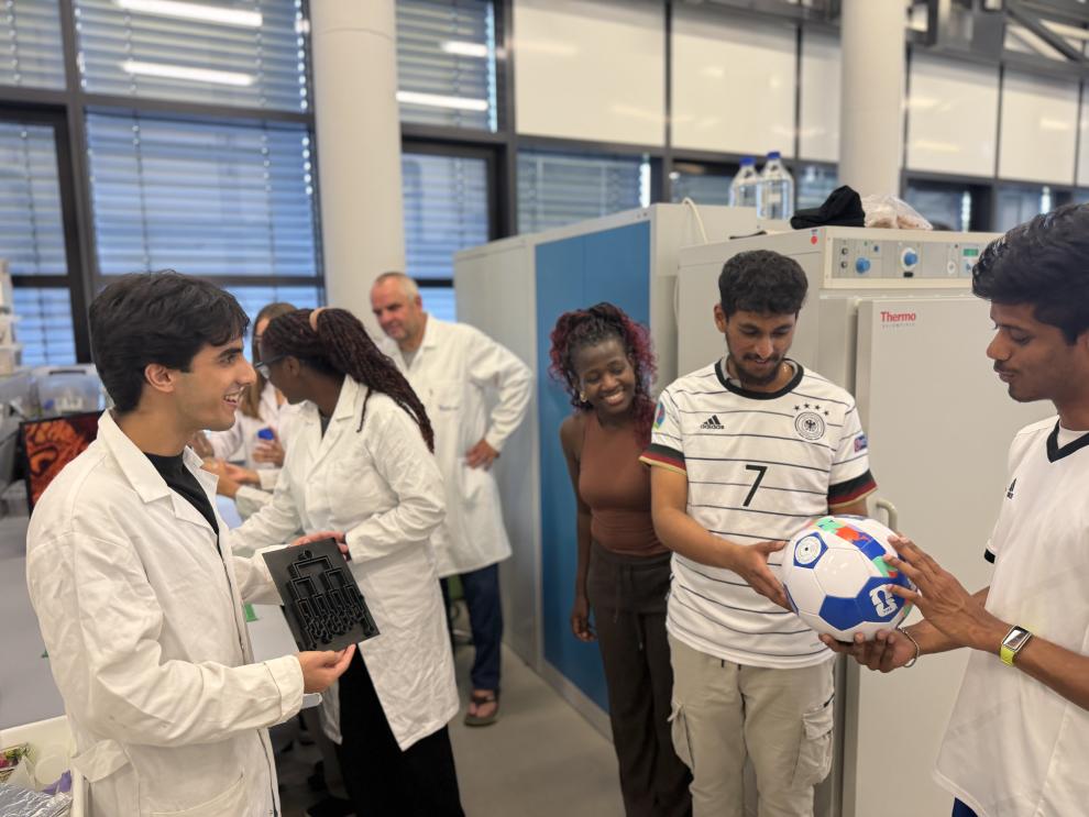 Eine Gruppe junger Leute unterhält sich fröhlich in einem Labor. Links hält ein lächelnder Mann im Laborkittel eine schwarze, 3D-gedruckte Nachbildung eines Turnierbaums. Rechts betrachten zwei Männer – einer davon im Deutschland-Trikot – gemeinsam einen Fußball. Im Hintergrund stehen weitere lächelnde Teammitglieder in Laborkitteln und Alltagskleidung.